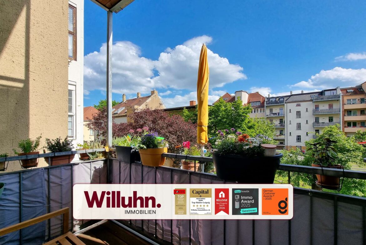 Balcony with potted plants and a tall closed yellow umbrella, city buildings across and blue sky; real estate banner on the railing reads Willuhn Immobilien.