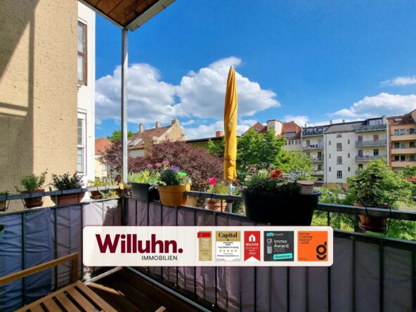 Balcony with potted plants and a tall closed yellow umbrella, city buildings across and blue sky; real estate banner on the railing reads Willuhn Immobilien.