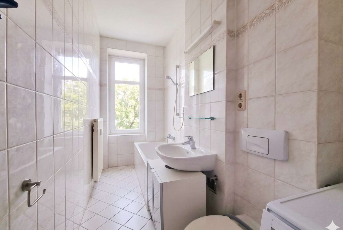 Narrow white-tiled bathroom with a window, sink, mirror, shower, and washing machine in the corner.