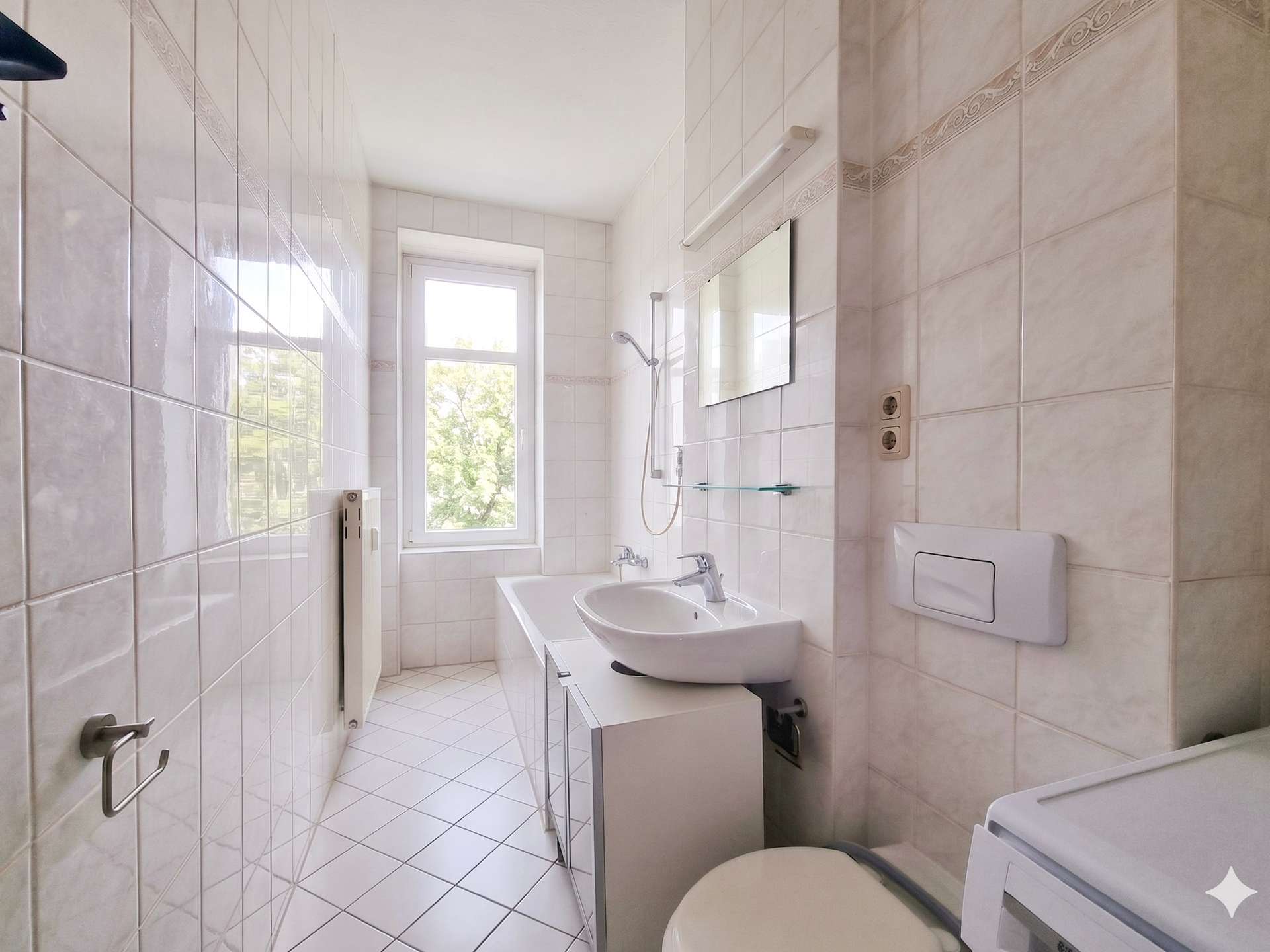 Narrow white-tiled bathroom with a window, sink, mirror, shower, and washing machine in the corner.