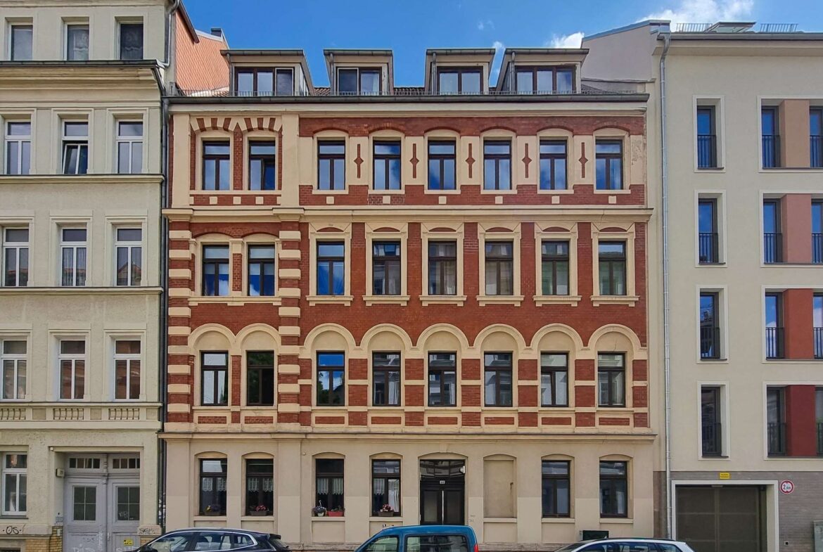 Facade of a historic red-brick building with beige trim and many windows, flanked by pale neighboring buildings under a blue sky.