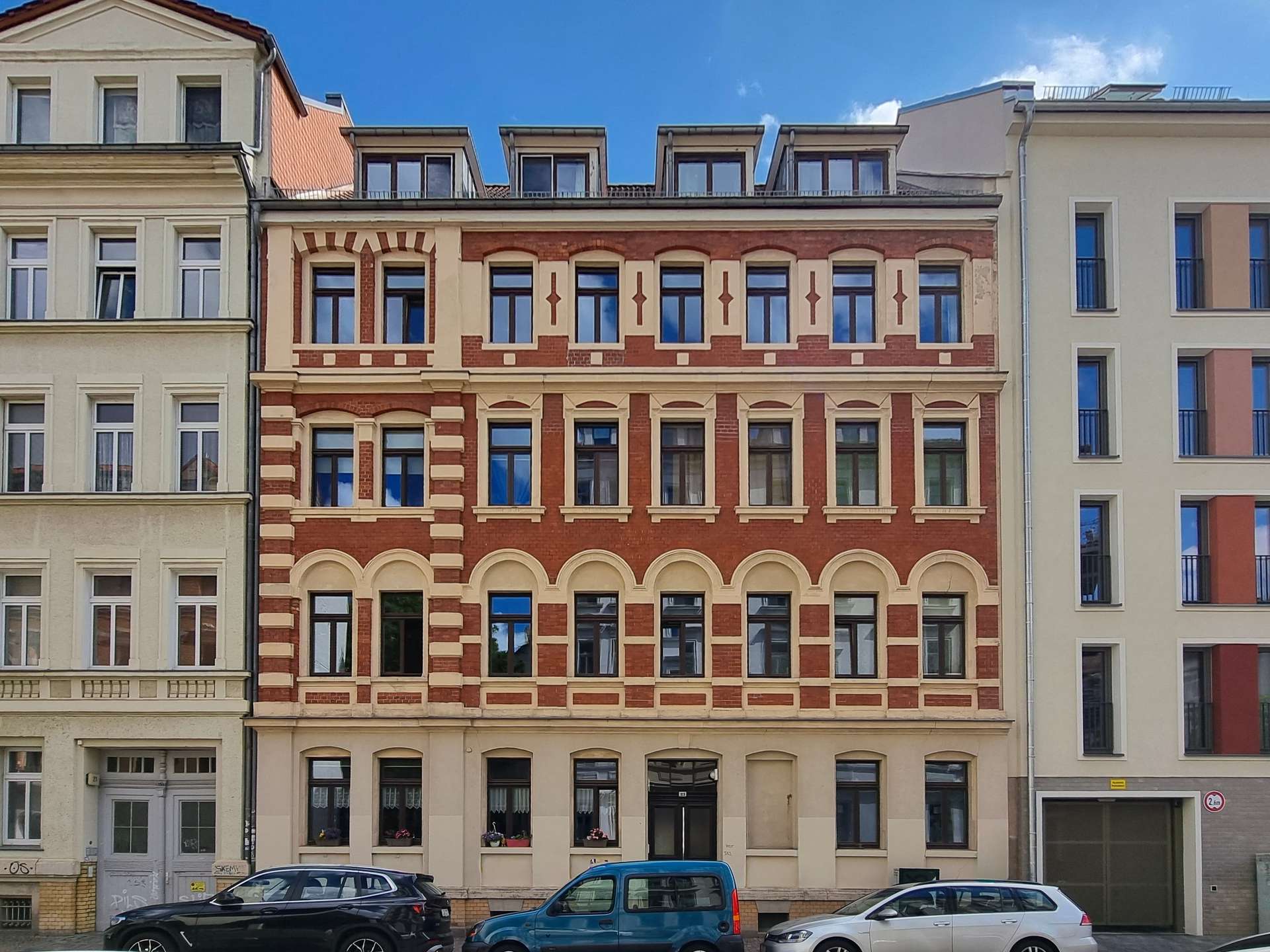 Facade of a historic red-brick building with beige trim and many windows, flanked by pale neighboring buildings under a blue sky.