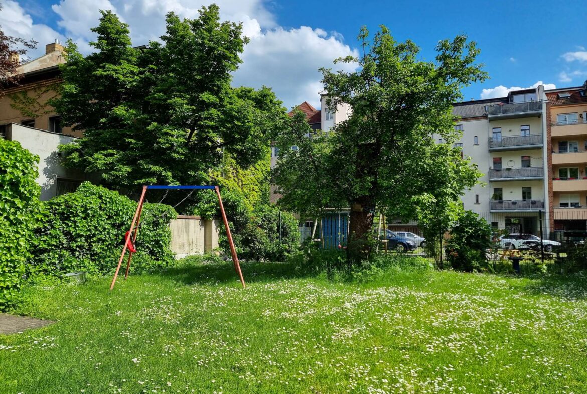 Green urban park with a swing set in the foreground, tall trees, and a row of apartment buildings against a blue sky with clouds.
