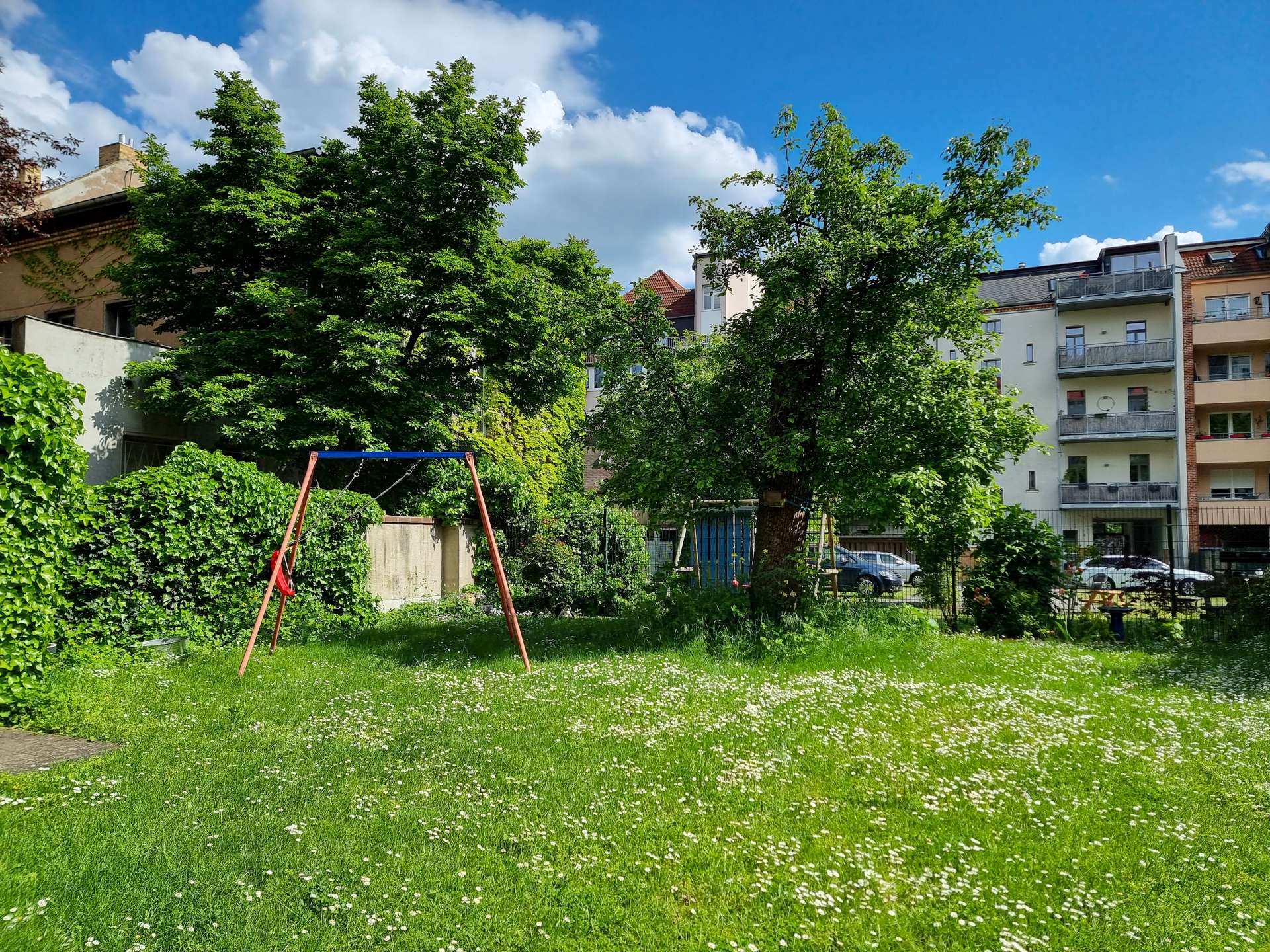 Green urban park with a swing set in the foreground, tall trees, and a row of apartment buildings against a blue sky with clouds.