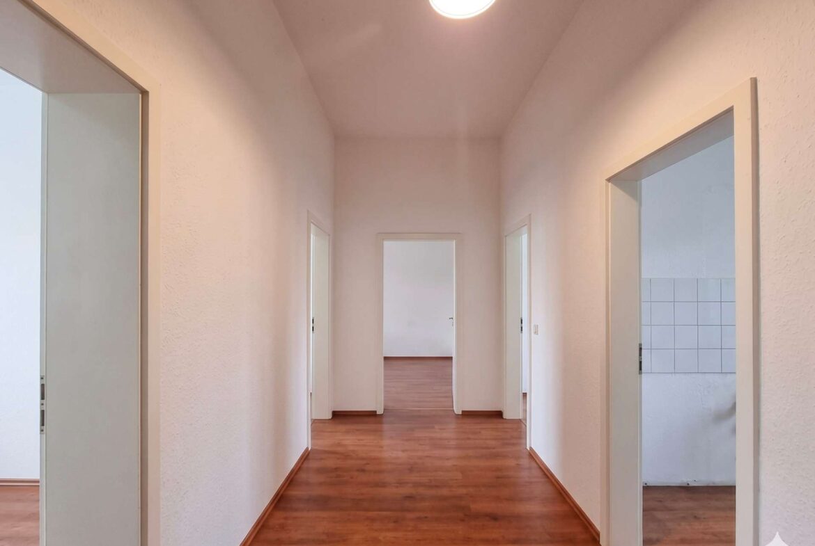 Long white hallway with wooden floors and multiple doorways on both sides, illuminated by a bright ceiling light.