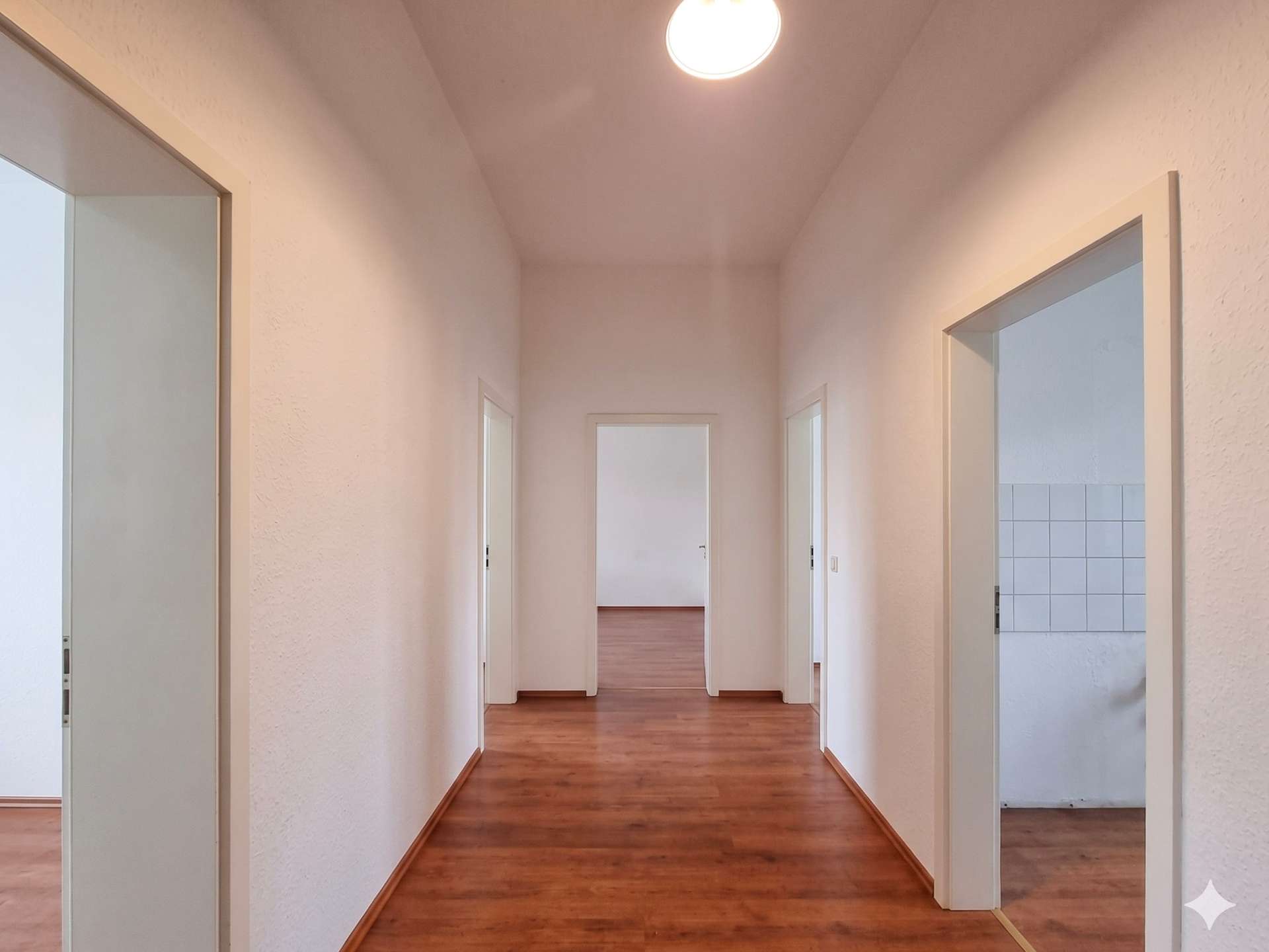 Long white hallway with wooden floors and multiple doorways on both sides, illuminated by a bright ceiling light.