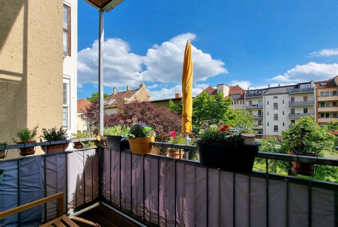 Balcony with potted plants and a folded yellow patio umbrella, overlooking apartment buildings against a blue sky.