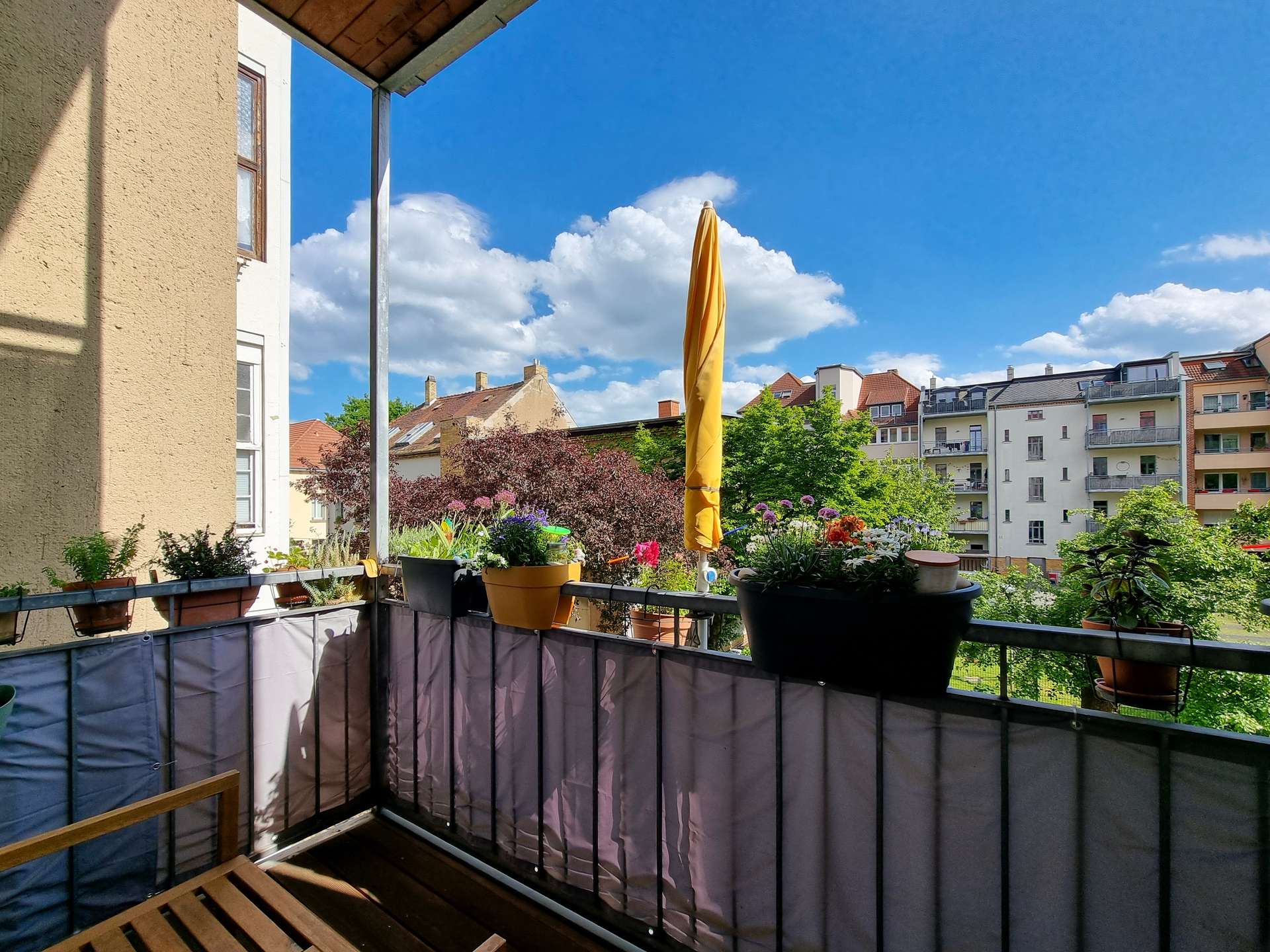Balcony with potted plants and a folded yellow patio umbrella, overlooking apartment buildings against a blue sky.