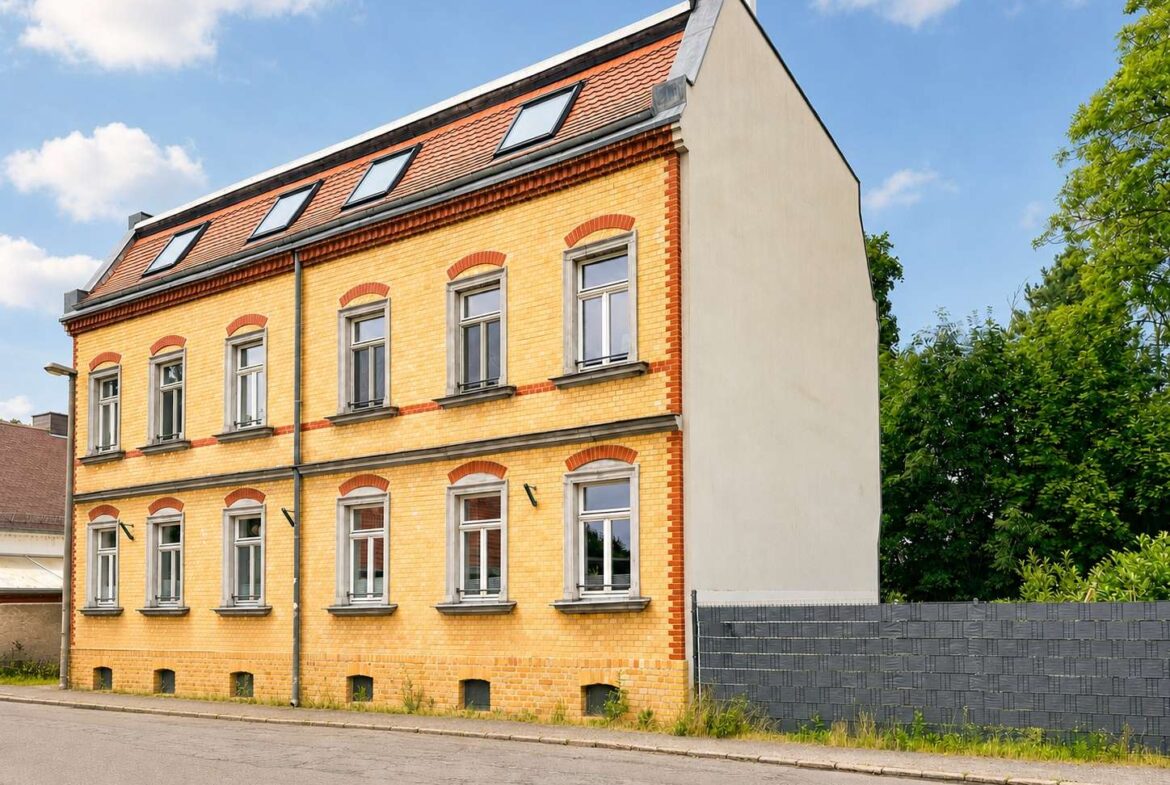 Three-story yellow brick building with red brick accents and arched window lintels on a quiet street, blue sky and green trees in the background