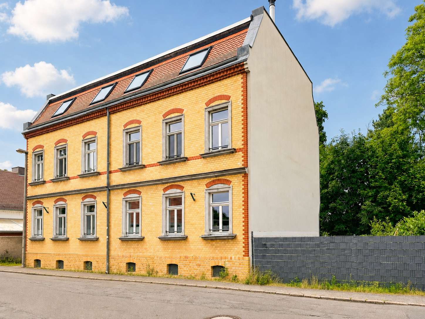 Three-story yellow brick building with red brick accents and arched window lintels on a quiet street, blue sky and green trees in the background