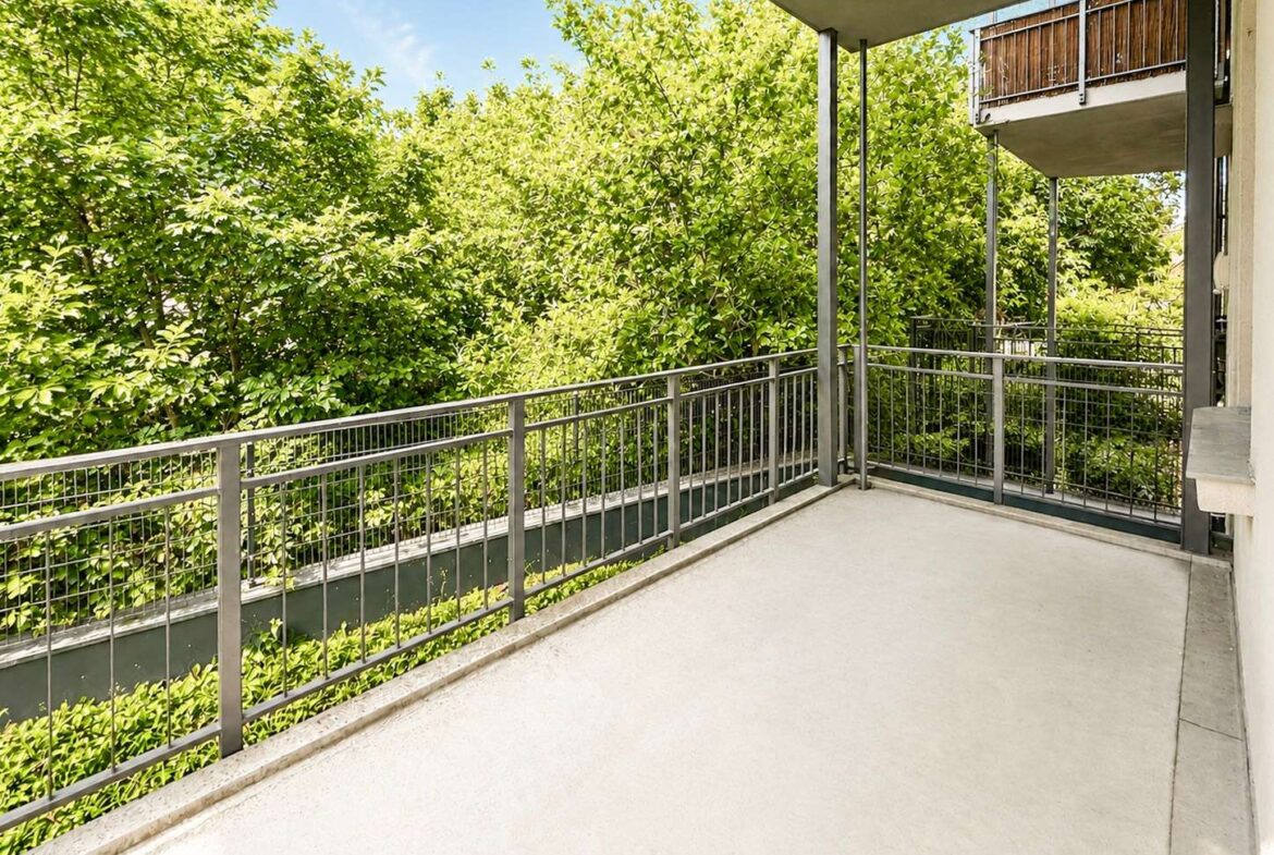 Balcony with gray metal railing overlooking dense green trees and blue sky; concrete floor and building support beams visible.
