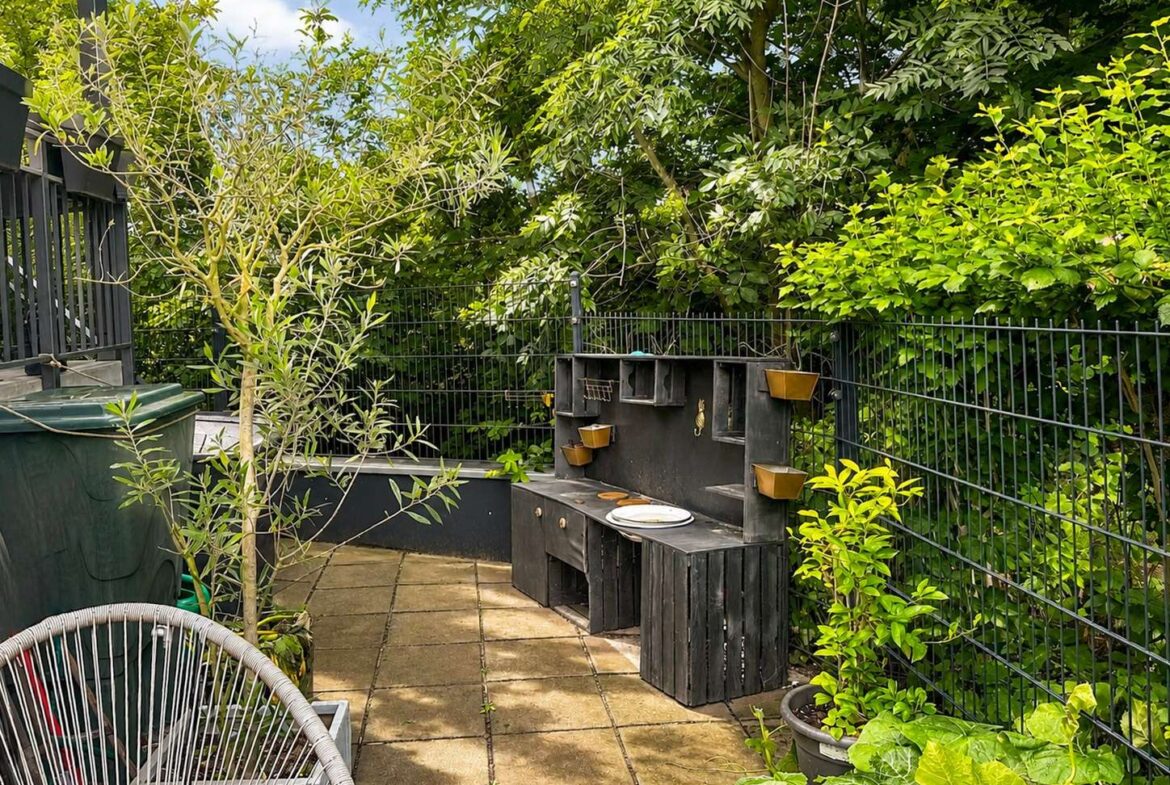 Backyard patio with a black wooden barbecue station along a metal fence, small planter boxes on the back wall, and lush greenery surrounding the area.