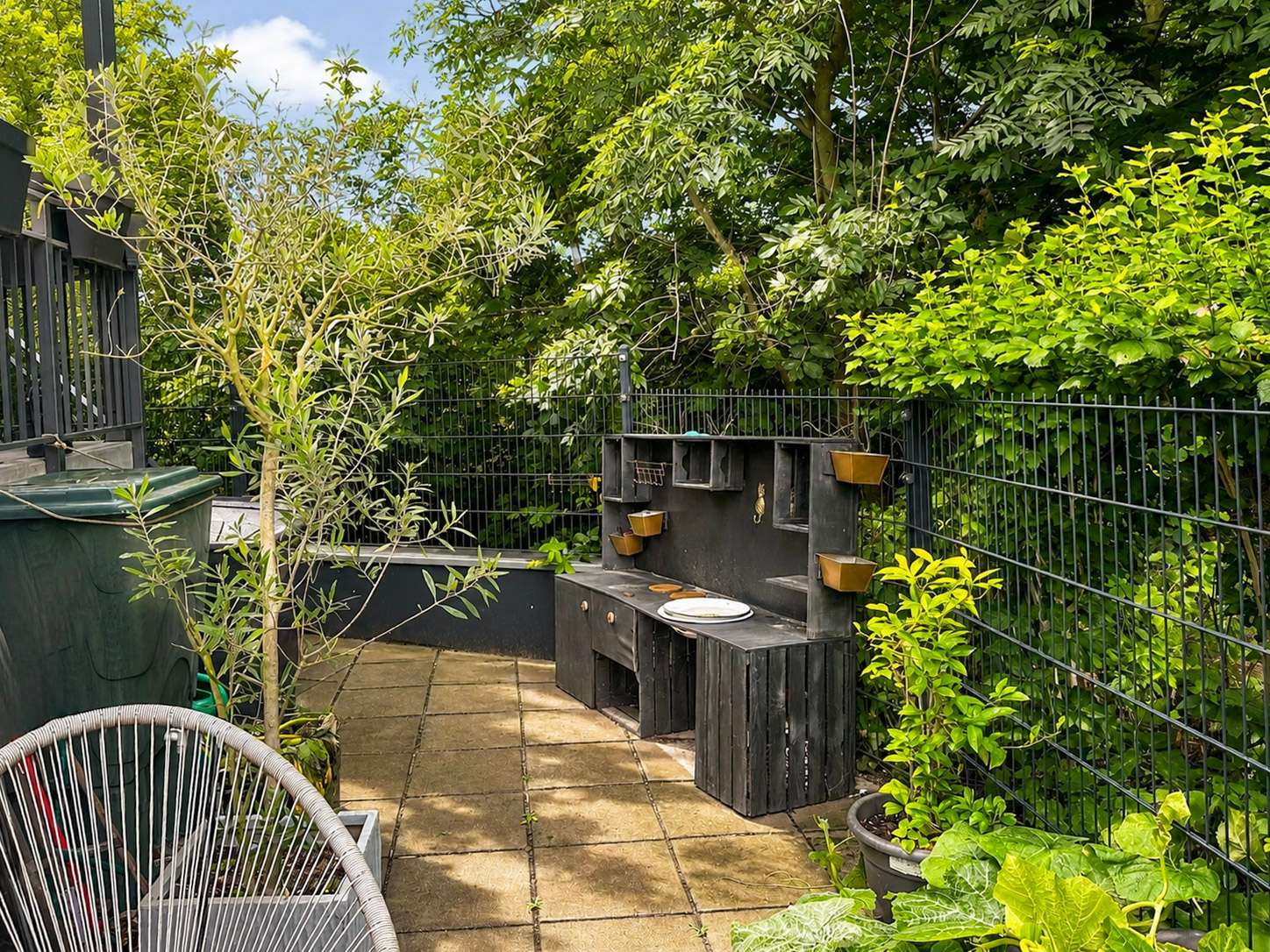 Backyard patio with a black wooden barbecue station along a metal fence, small planter boxes on the back wall, and lush greenery surrounding the area.