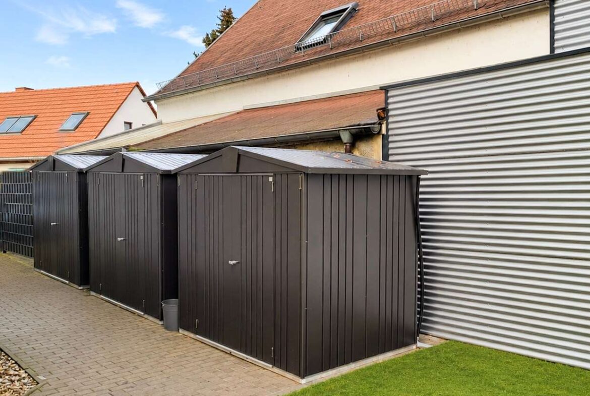 Row of black metal storage sheds along a paved walkway beside a beige building with a red-tile roof.
