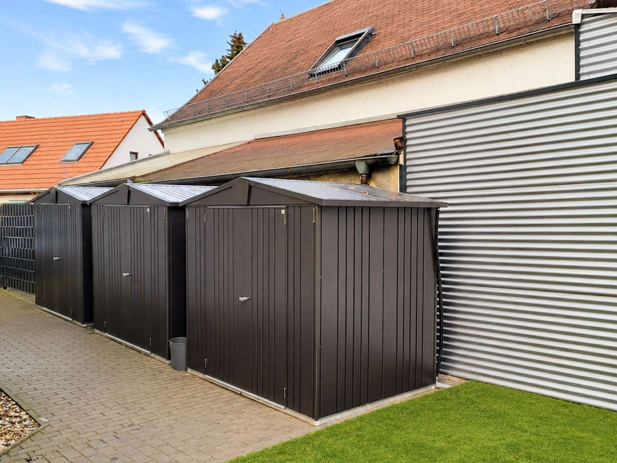 Row of black metal storage sheds along a paved walkway beside a beige building with a red-tile roof.