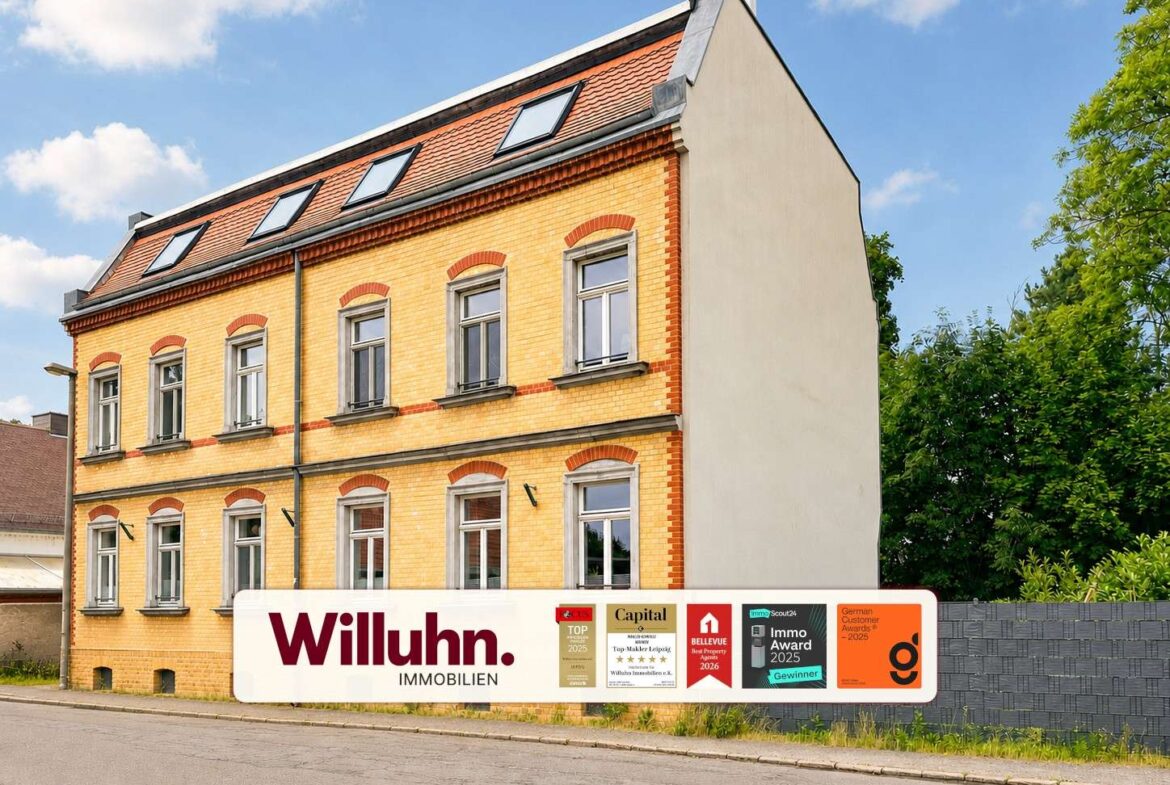 Three-story yellow brick building with red brick trim and arched window tops, beside a sign reading 'Willuhn Immobilien' and award logos on the sidewalk banner.