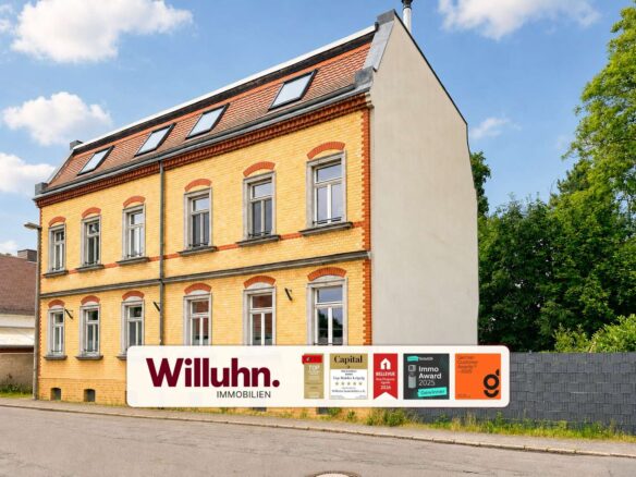 Three-story yellow brick building with red brick trim and arched window tops, beside a sign reading 'Willuhn Immobilien' and award logos on the sidewalk banner.