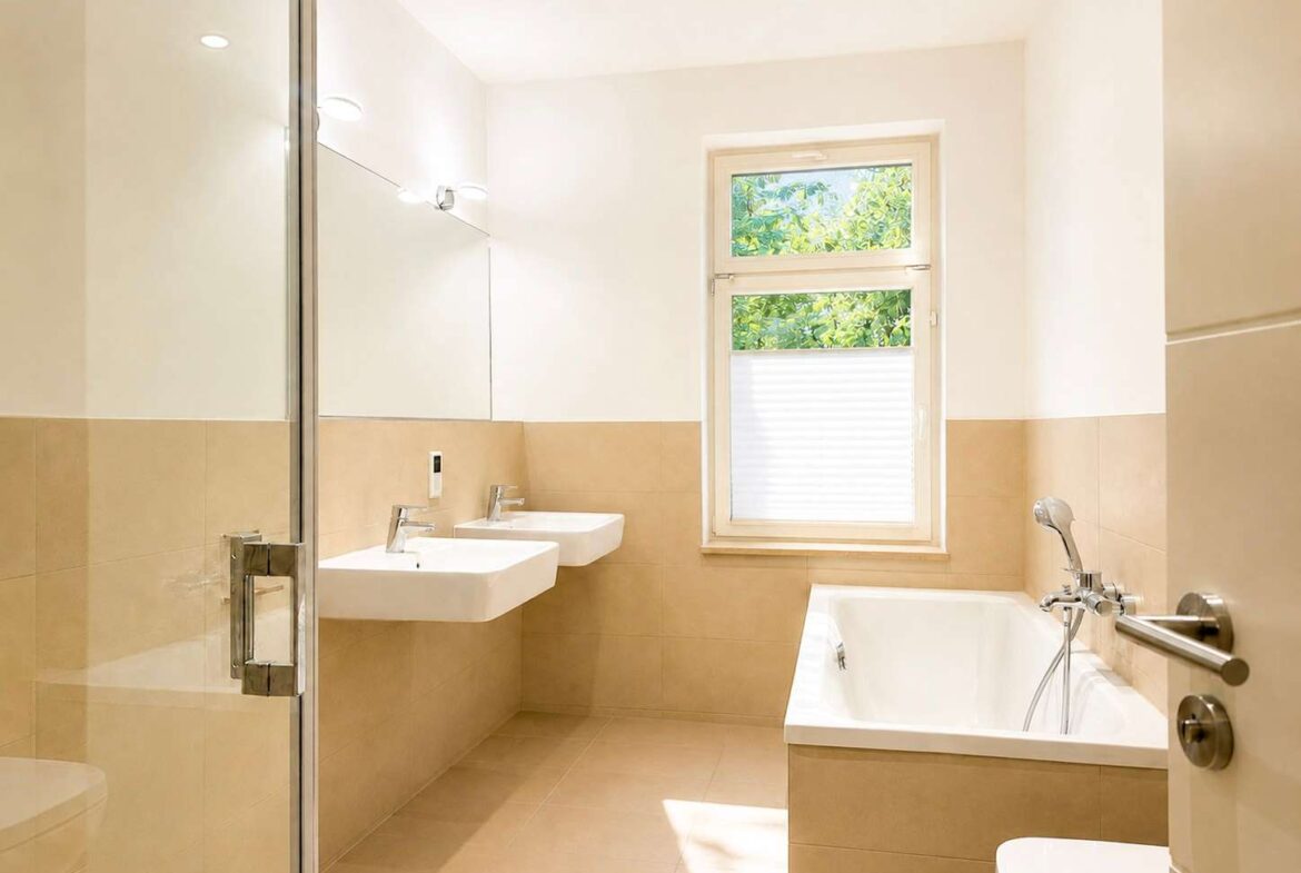 Bright modern bathroom with two wall-mounted sinks, large mirror, and glass shower enclosure on beige tile walls and floor.