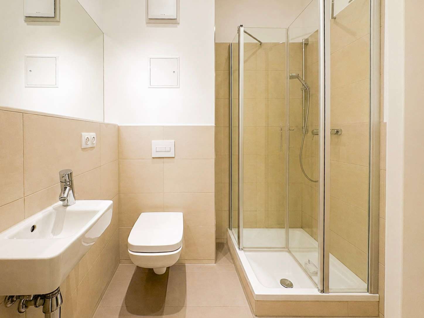 Modern beige-tiled bathroom with a wall-mounted sink, wall switch, toilet, and glass-enclosed shower.