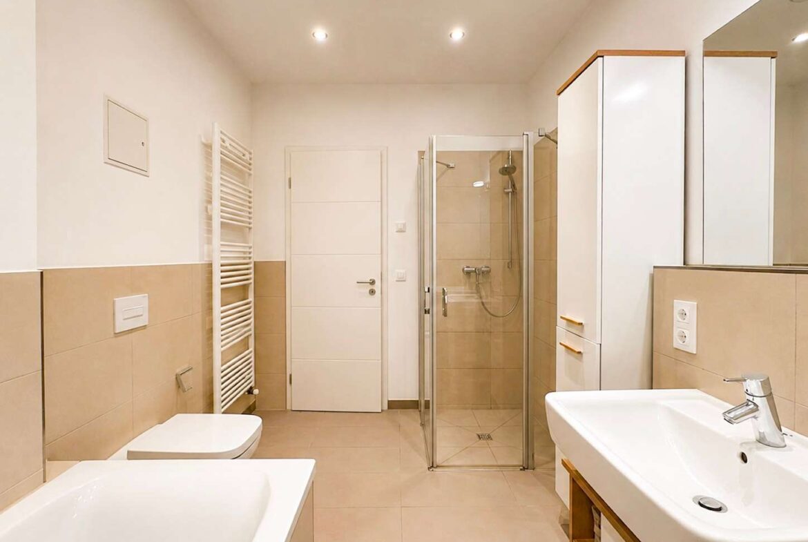Bright, modern bathroom with a glass shower enclosure, white sink, and beige tiled walls and floor; bathtub visible on the left and a heated towel rail along the wall.
