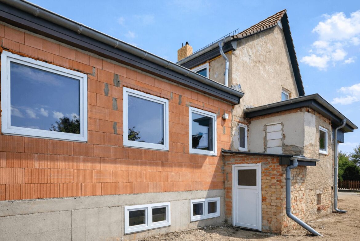 Two-story house under renovation with orange brick wall, exposed plaster, and white-framed windows against a blue sky.