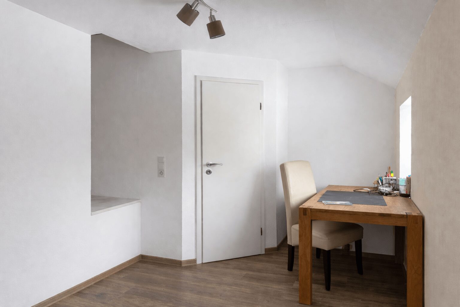 Minimalist home office corner with a wooden desk, beige chair, and a white door to the left.