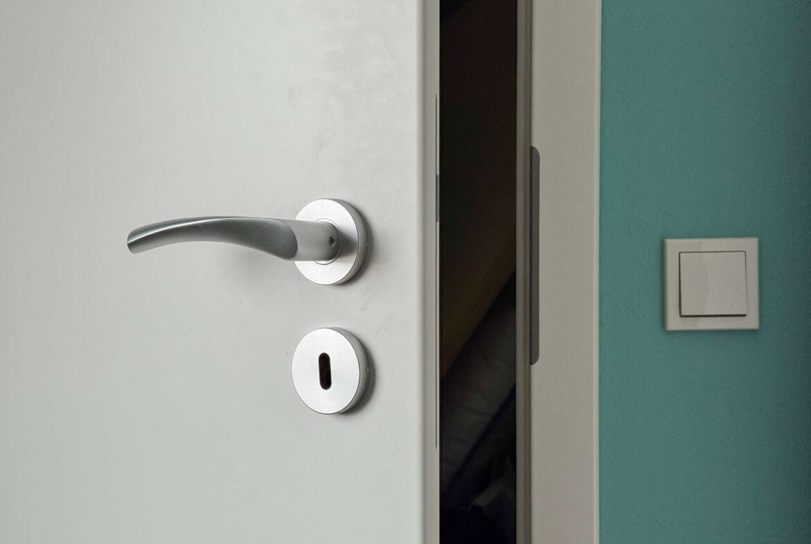 White door with a silver lever handle and deadbolt, slightly ajar to a dark interior; teal wall with a light switch on the right.