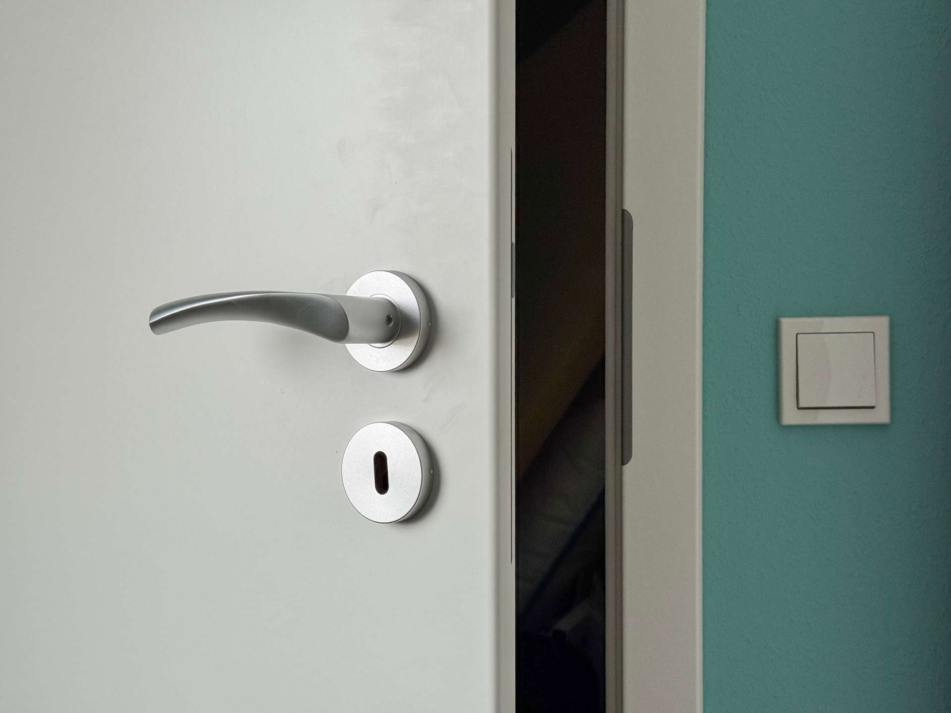 White door with a silver lever handle and deadbolt, slightly ajar to a dark interior; teal wall with a light switch on the right.