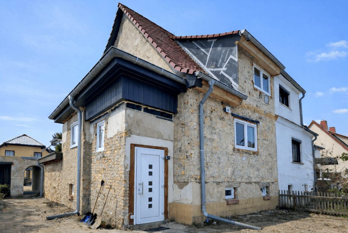 Stone two-story house under renovation with exposed stone walls, white front door, and gray gutter system against a clear blue sky