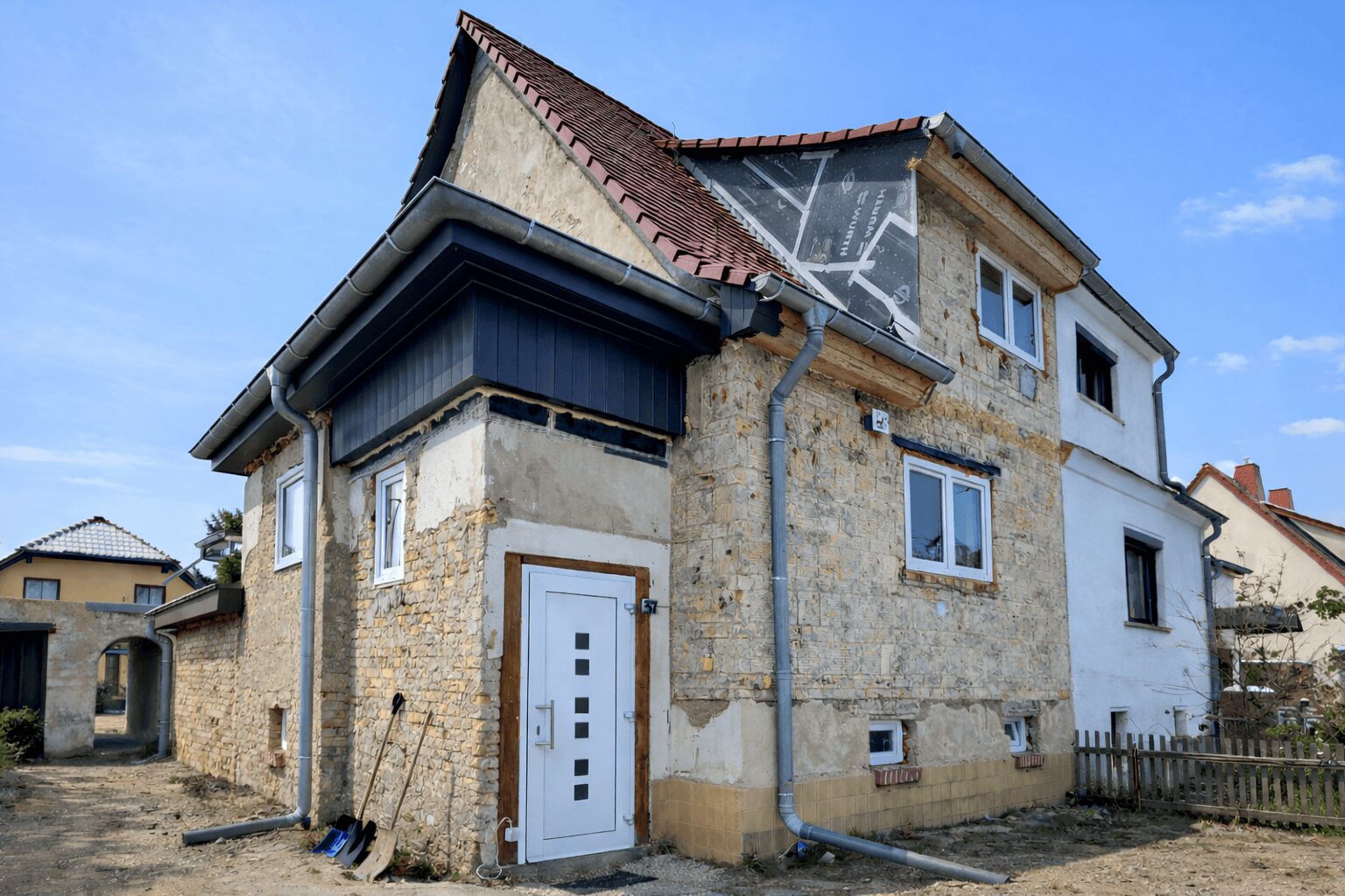 Stone two-story house under renovation with exposed stone walls, white front door, and gray gutter system against a clear blue sky