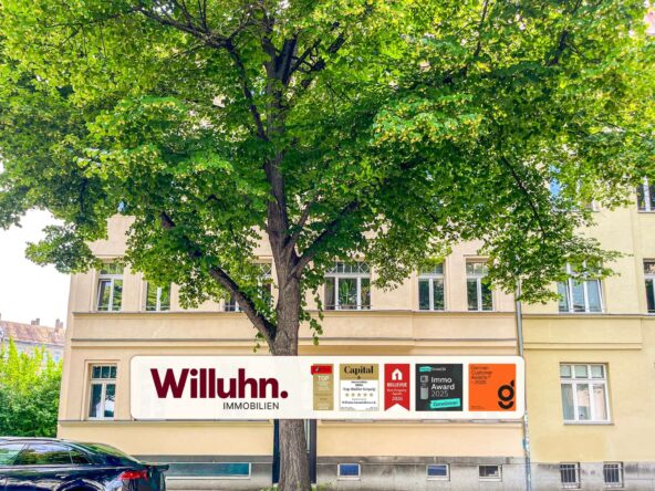 Tree in front of a beige building with a 'Willuhn Immobilien' sign and award plaques beneath it.