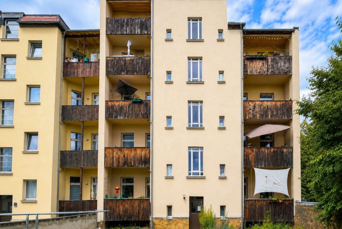 Beige apartment building with multiple wooden balconies and small garden in front.