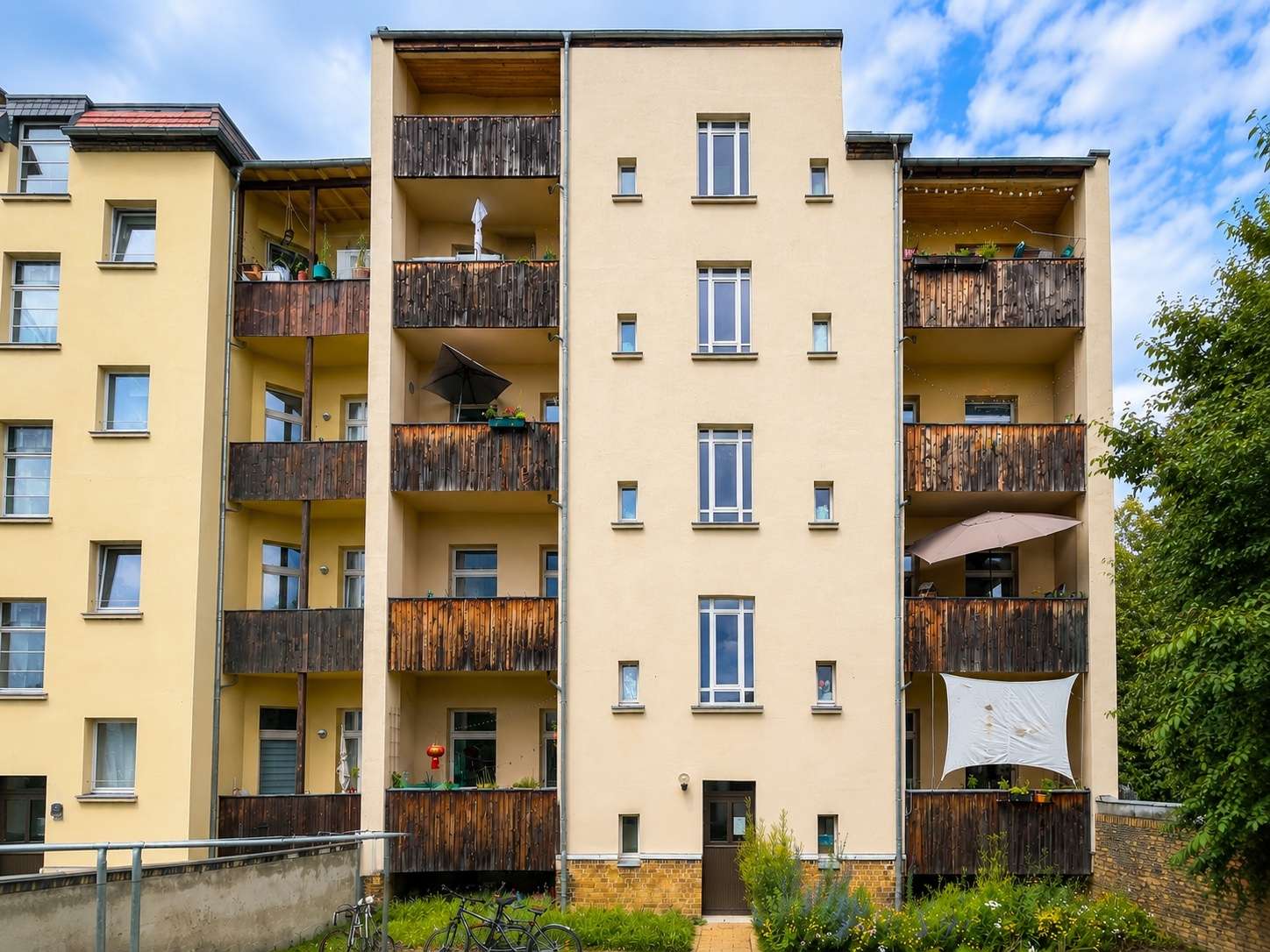 Beige apartment building with multiple wooden balconies and small garden in front.