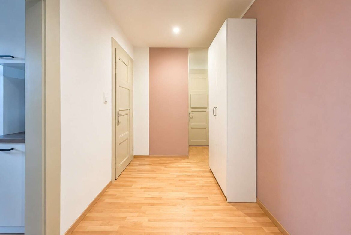 Bright hallway with light wood floor, white walls and a pink accent wall; a tall white wardrobe on the right and a door at the end of the corridor.