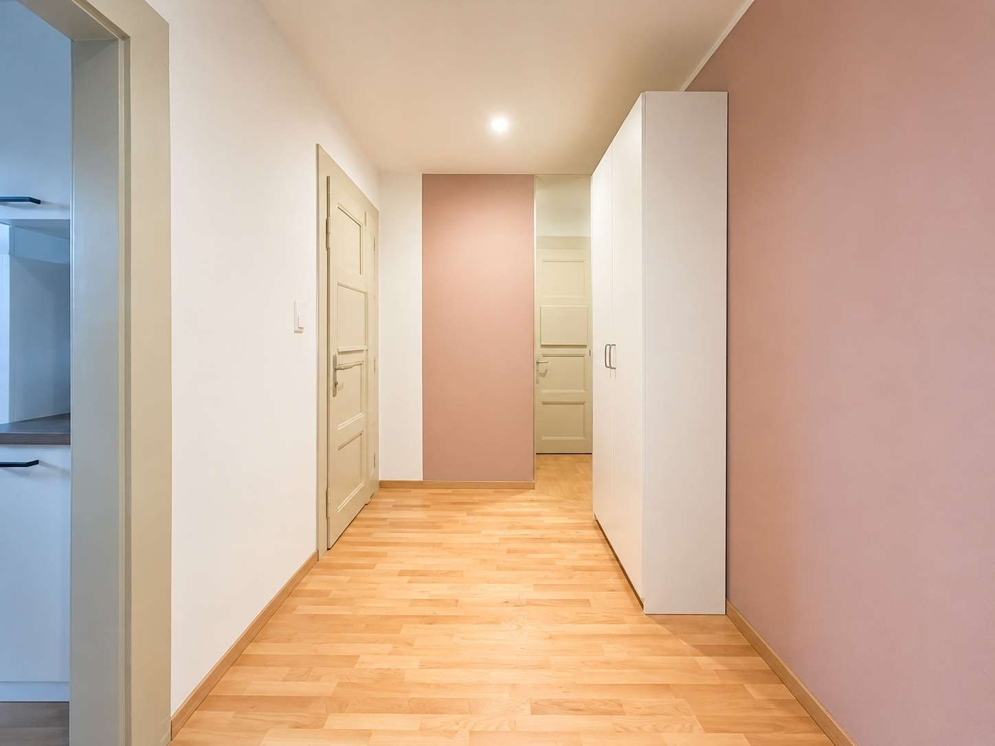 Bright hallway with light wood floor, white walls and a pink accent wall; a tall white wardrobe on the right and a door at the end of the corridor.