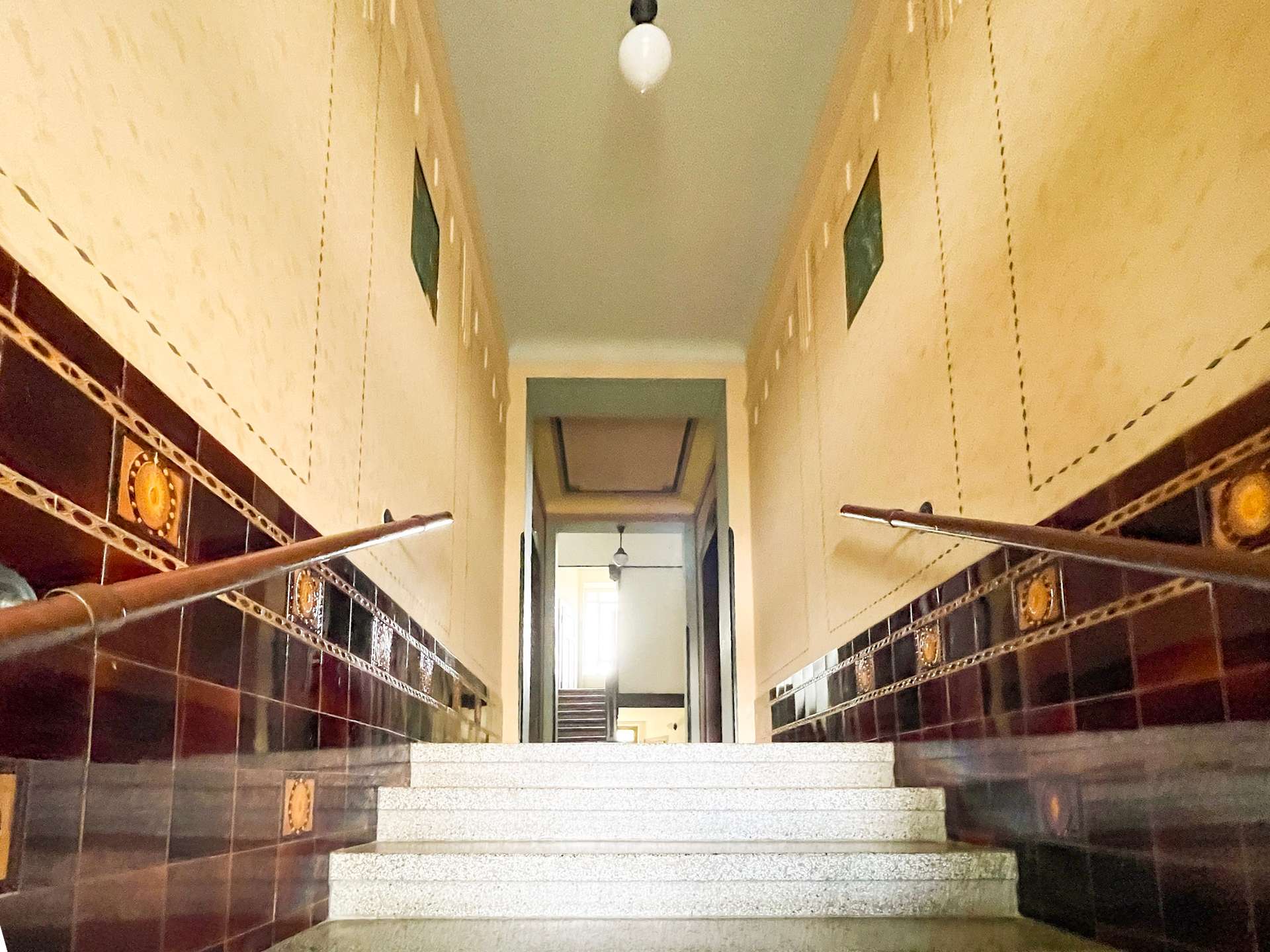 Long view of a tiled stairwell with brown glossy wall tiles and decorative round inserts, converging to a doorway at the top.