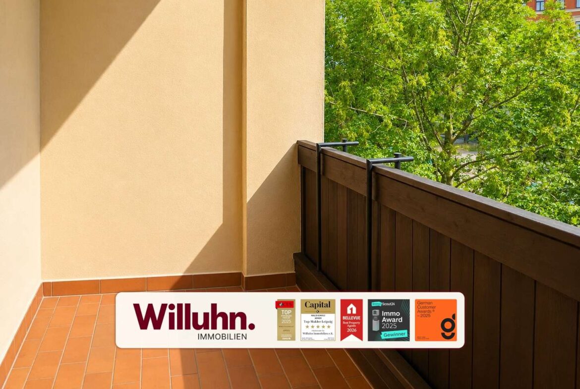 Sunlit balcony with terracotta tiles and beige walls, brown railing, green tree in the background, and a Willuhn Immobilien banner along the bottom edge.