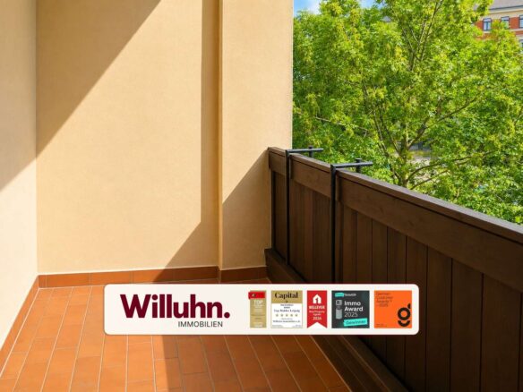 Sunlit balcony with terracotta tiles and beige walls, brown railing, green tree in the background, and a Willuhn Immobilien banner along the bottom edge.