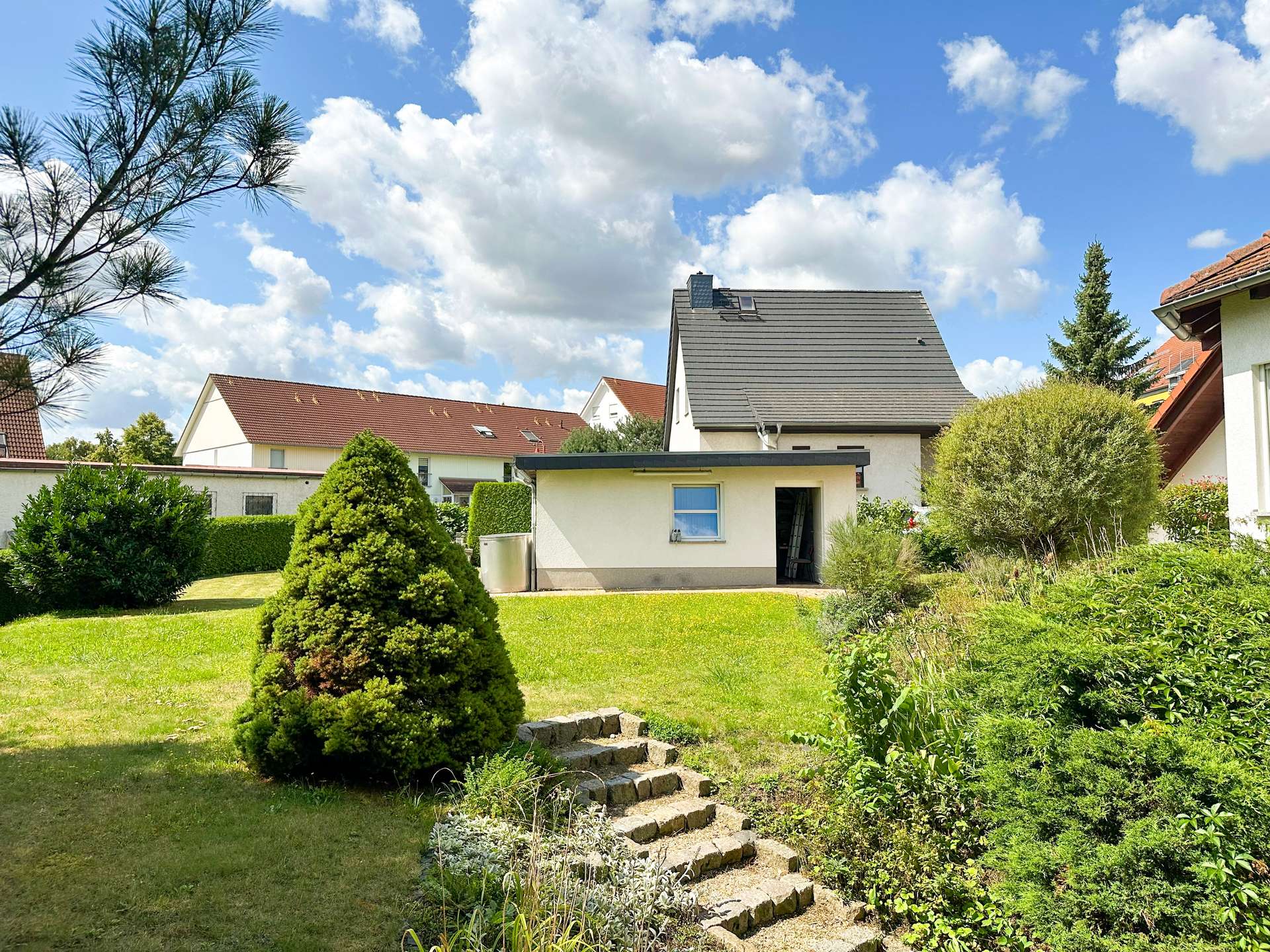 Suburban backyard with a white house and steep gray roof, trimmed bushes, and stone steps leading toward the yard under a blue sky with clouds.