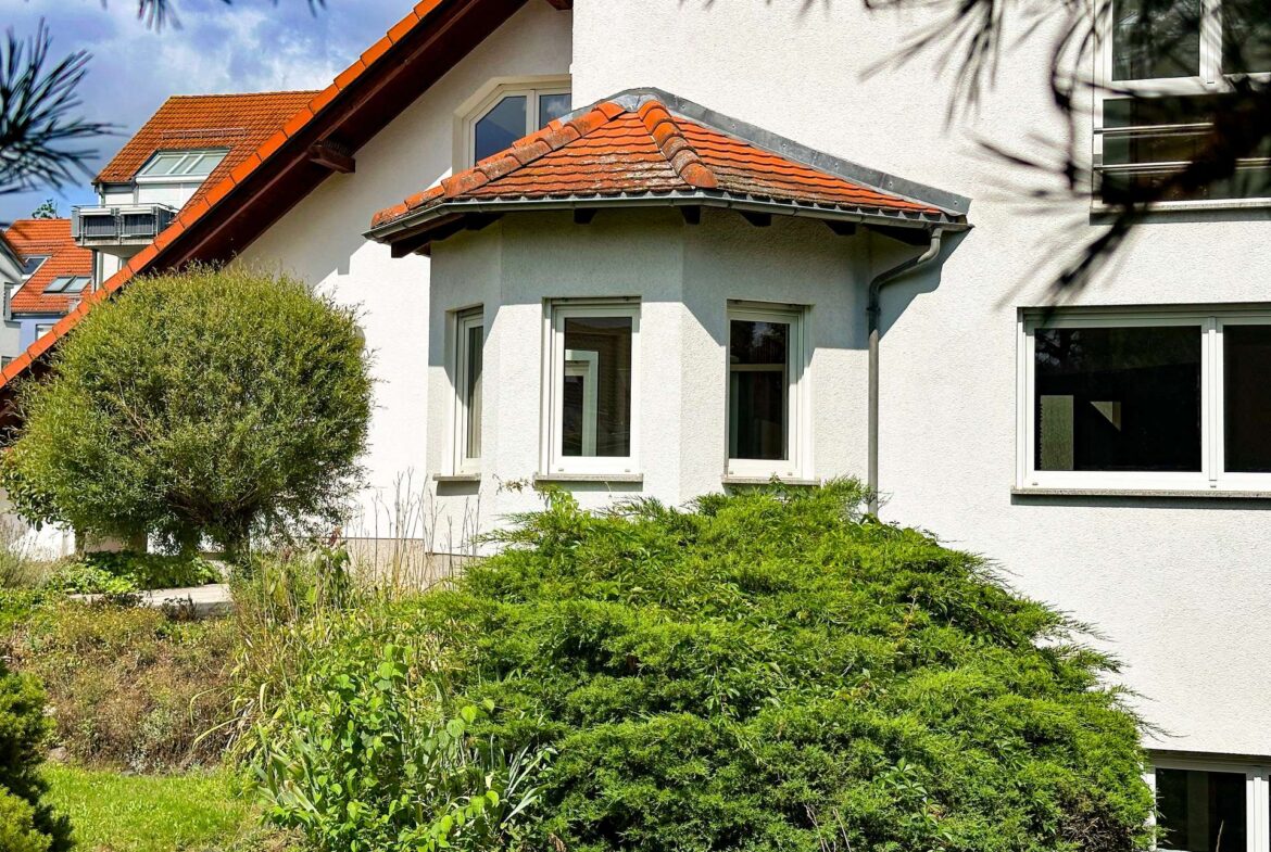 White stucco house with red tiled roof and large bay window, surrounded by dense green shrubs and garden vegetation.