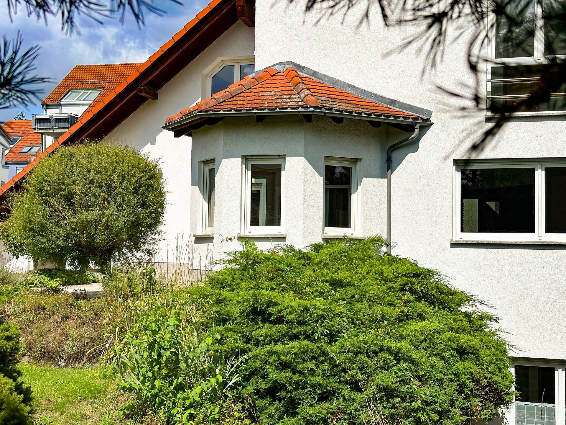 White stucco house with red tiled roof and large bay window, surrounded by dense green shrubs and garden vegetation.