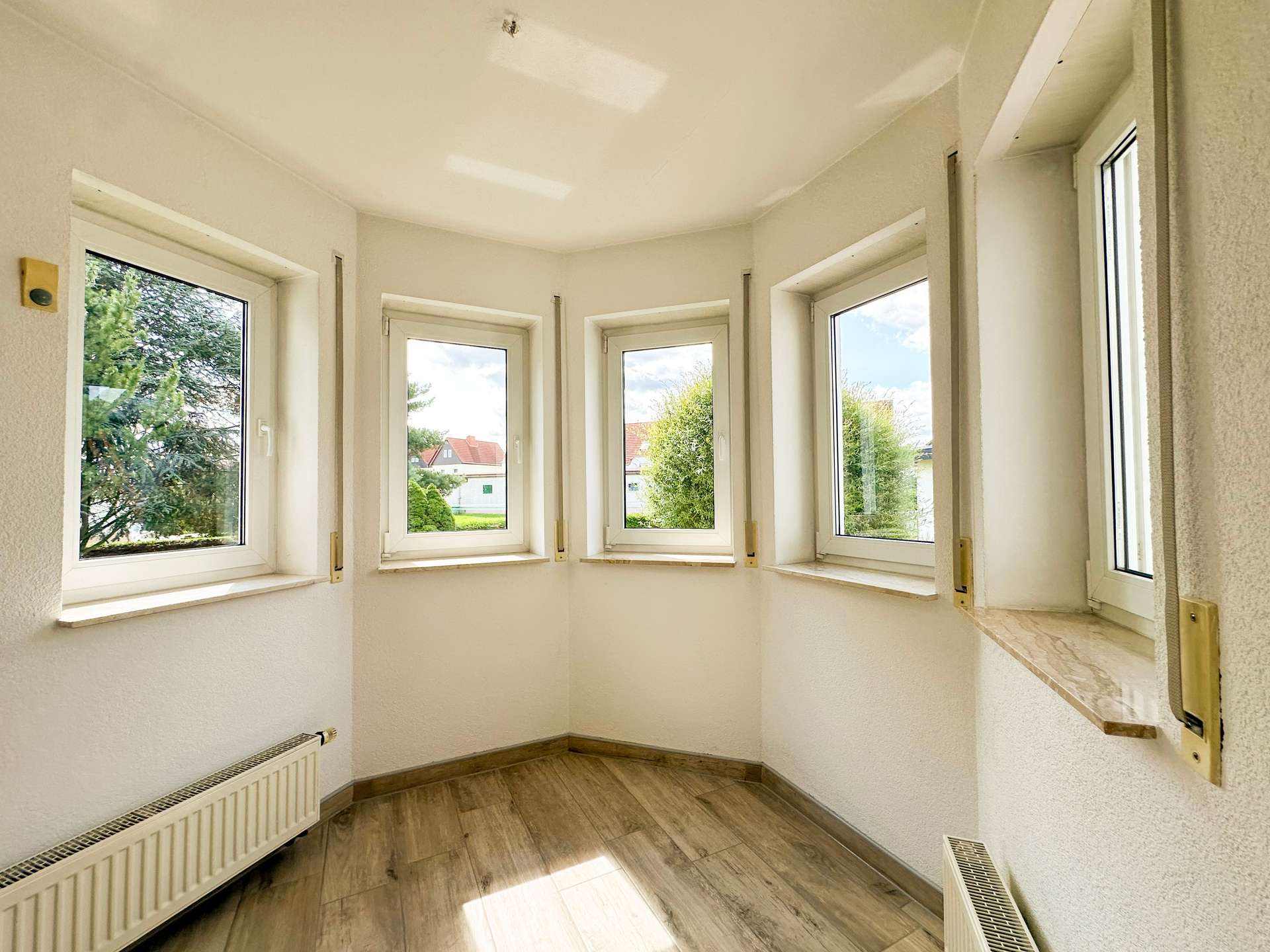 Bright corner room with a curved wall of windows, white walls, and a wooden floor; radiators along the lower walls.