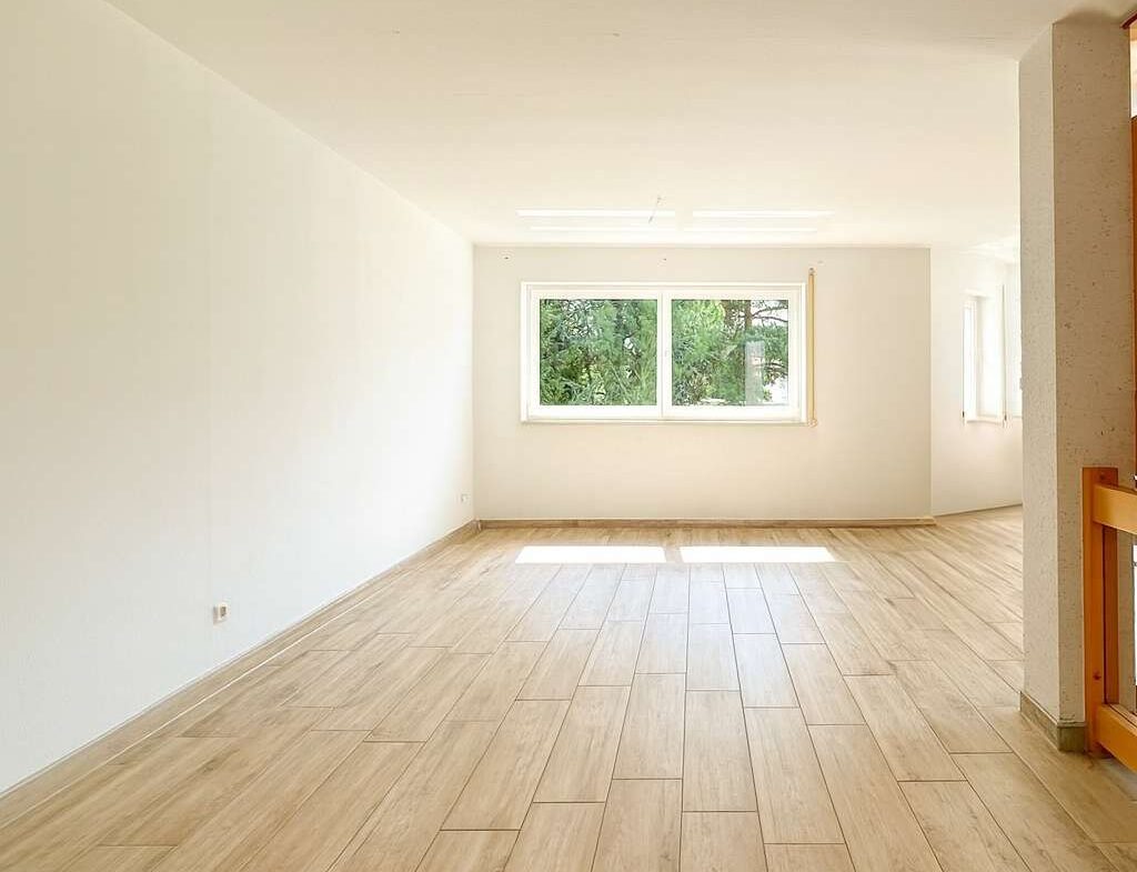 Empty bright living room with light wood flooring and a large window letting in natural light