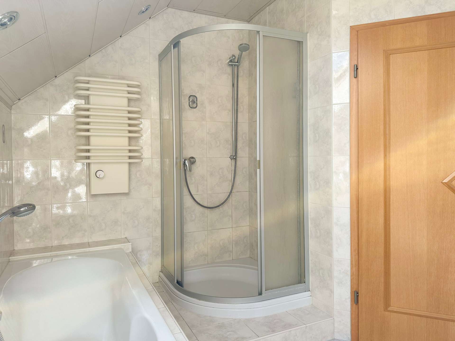 Curved corner shower enclosure with glass doors in a tiled bathroom, next to a white heated towel radiator and a wooden door fixtures visible nearby.
