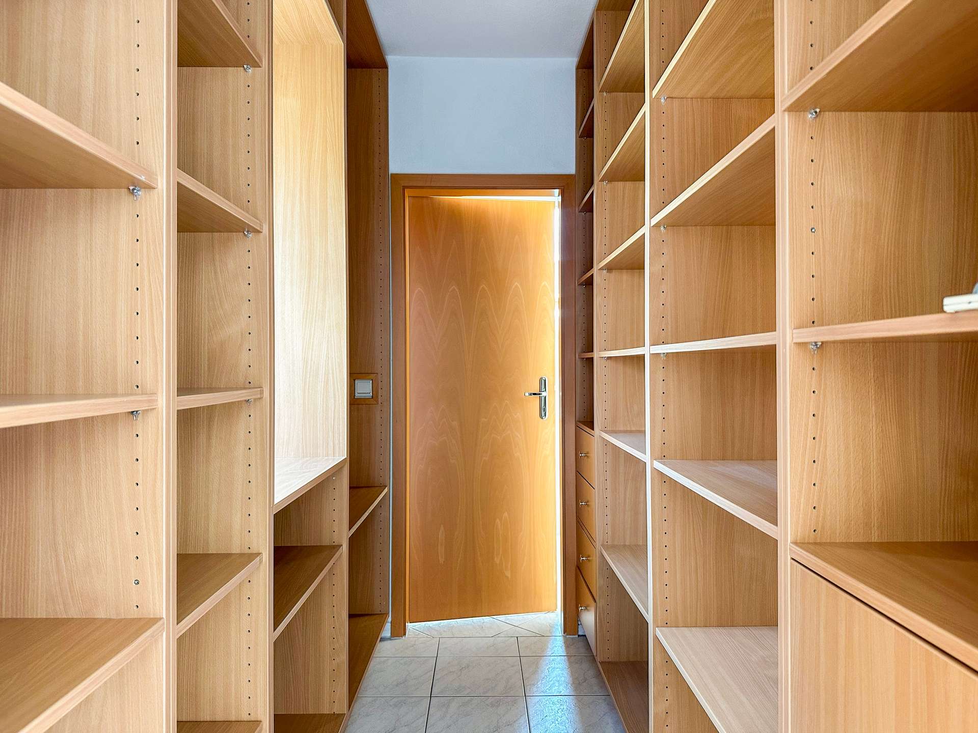 Wooden walk-in closet with tall, adjustable shelves on both sides and a door at the far end.