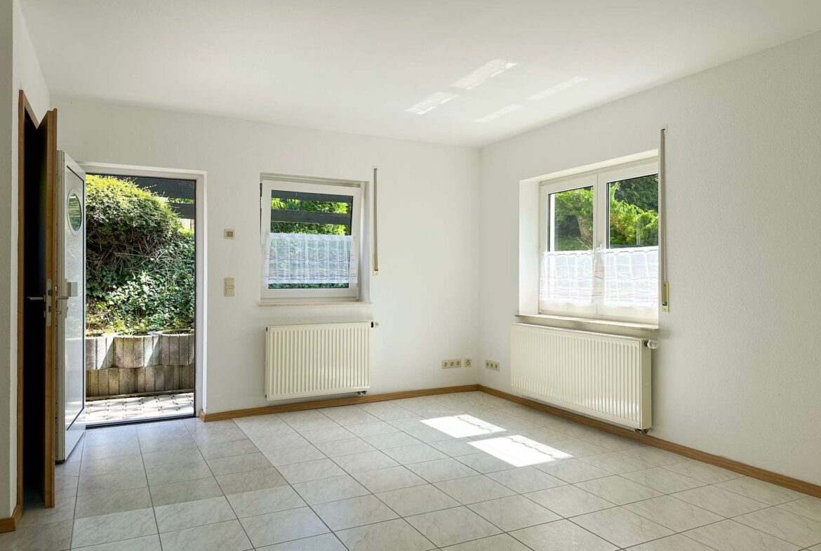 Bright empty room with white walls, tiled floor, and two radiators; open door to a garden view outside.