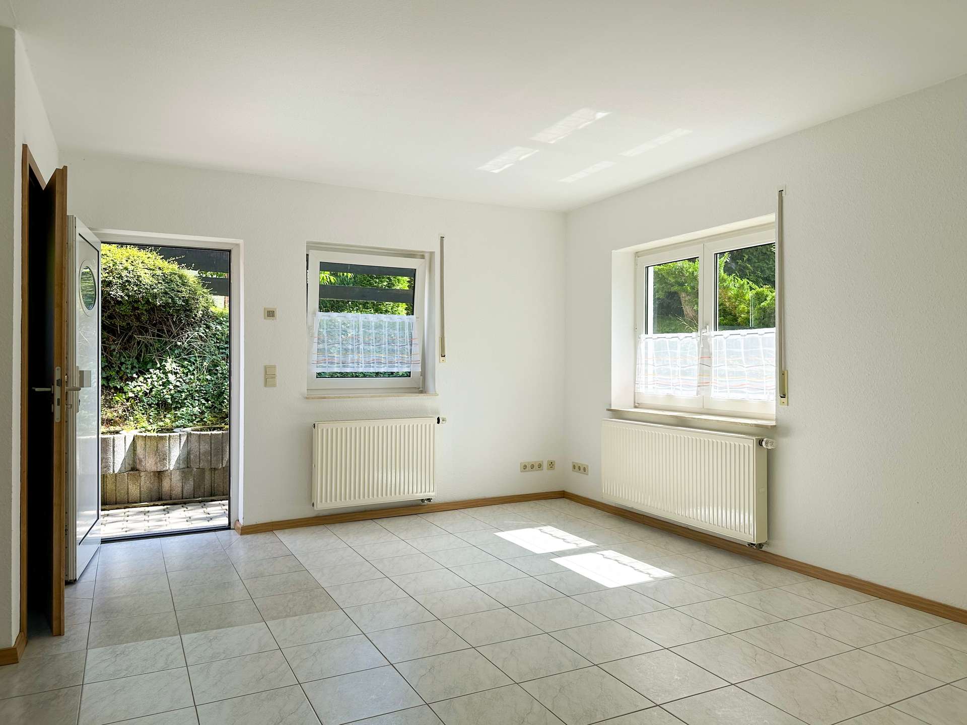 Bright empty room with white walls, tiled floor, and two radiators; open door to a garden view outside.