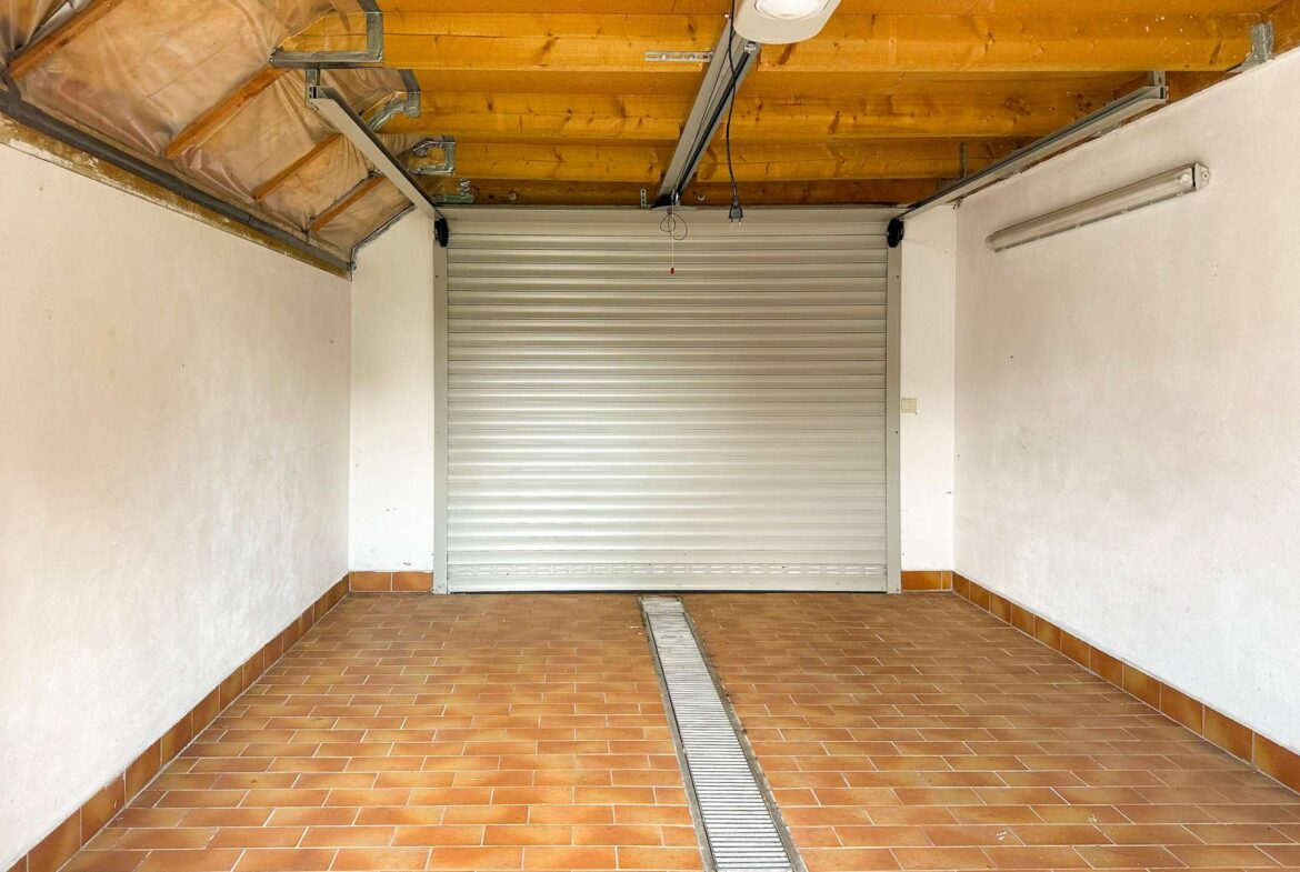 Garage interior with a closed metal roll-up door, exposed wooden ceiling beams, and a central floor drain.