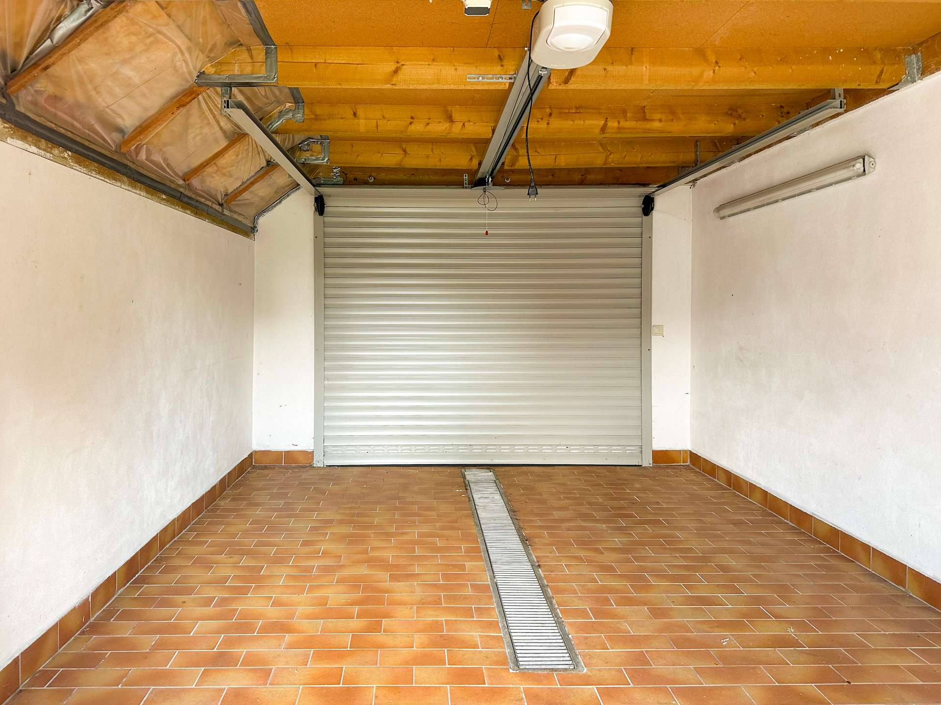 Garage interior with a closed metal roll-up door, exposed wooden ceiling beams, and a central floor drain.