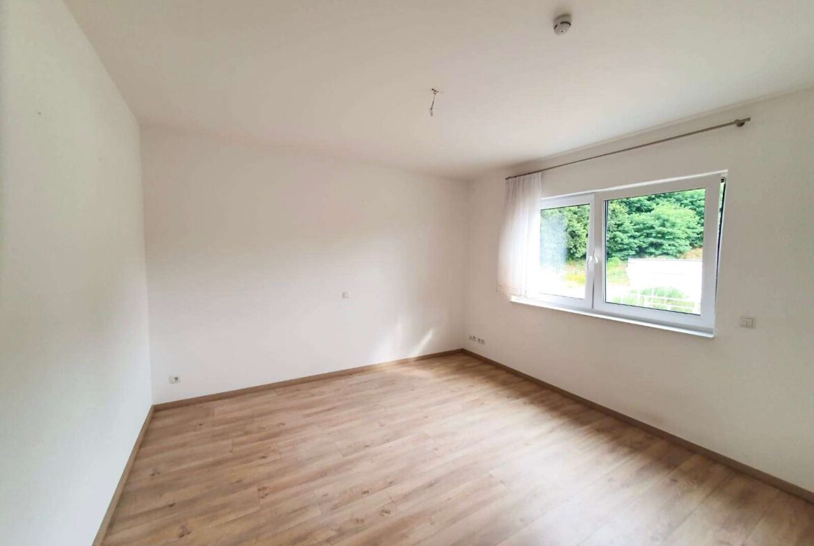 Empty room with white walls and light wood flooring, large window with sheer curtains letting in daylight.